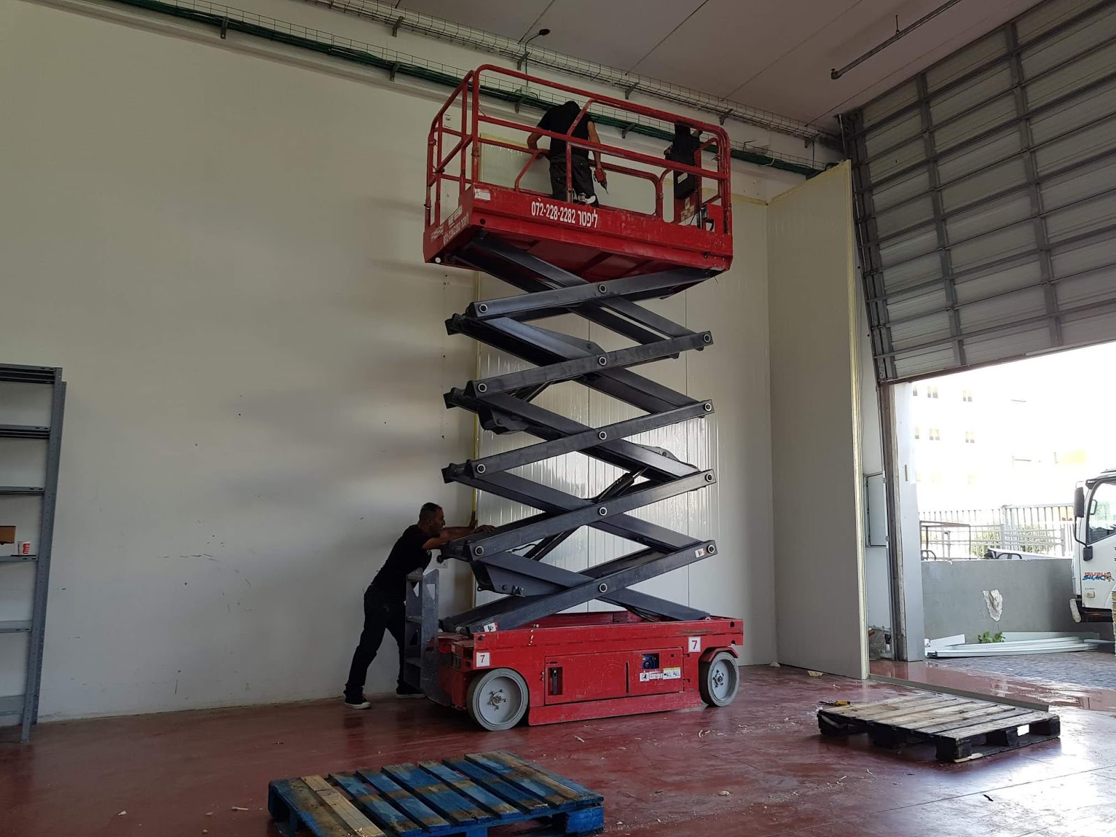 במת הרמה מספריים אדומה נמצאת בחלל עם שני אנשים העובדים עליה ולידה, ולידה פתח גדול.